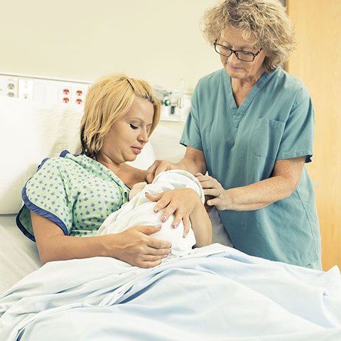 Im Krankenhaus-Zimmer: Hebamme steht bei Mutter und Baby am Bett. 